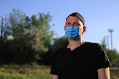 Günlük giysiler ve tıbbi maske takmış beyaz bir genç ya da yetişkin bir adam şehir parkında dinleniyor..