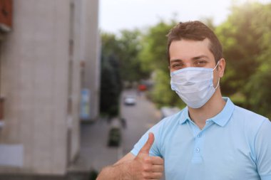 Tıbbi maske takmış, şehir ortamının bulanık arka planında yüzünü koruyan beyaz bir adamın yaşam tarzı portresi.