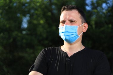 Günlük giysiler ve tıbbi maske takmış beyaz bir genç ya da yetişkin bir adam şehir parkında dinleniyor..