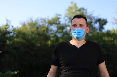 Günlük giysiler ve tıbbi maske takmış beyaz bir genç ya da yetişkin bir adam şehir parkında dinleniyor..