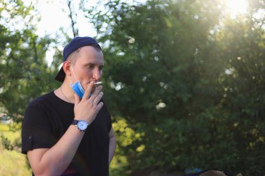 Genç beyaz bir adam ya da genç bir beyaz adam çenesinde tıbbi bir maskeyle sigara içiyor..