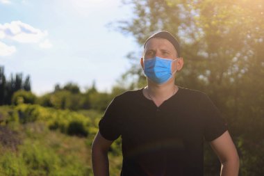 Günlük giysiler ve tıbbi maske takan beyaz bir genç ya da yetişkin adam şehir dışında doğanın tadını çıkarıyor. Ayarlanmış arkaplan
