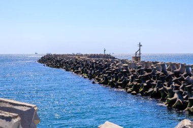 Güneş ışığıyla aydınlanan okyanusta küçük tekneleri olan bir dalgakıranda beton tetrapodlar.