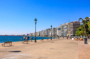 İnsanların banklarda yürüyüp oturduğu bir plaj sahnesi. Sahil yüksek binaları olan bir şehre yakındır. 8 Ağustos 2024 Selanik Yunanistan