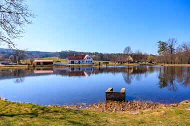 Sakin kırsal göl manzarası. Evlerin yansımaları ve açık mavi gökyüzü. 21 Mart 2025, Holasovice, Çek.