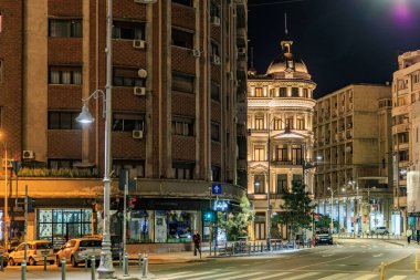 Sakin bir caddede aydınlatılmış tarihi ve modern binaların olduğu gece manzarası. 8 Mart 2025 Bükreş Romanya.