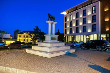 Modern binanın yakınındaki şehir meydanında gece vakti aydınlatılmış Roma kurt heykeli. 3 Mayıs 2025 Romanya.