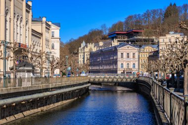 Açık mavi gökyüzünün altında tarihi binalar ve nehirler olan manzaralı şehir manzarası. 19 Mart 2025, Karlovy Vary, Çek Cumhuriyeti.