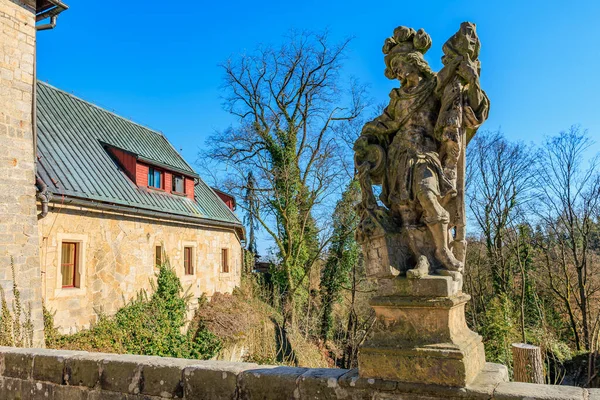 Tarihsel taş heykel ve kırsal yapı açık mavi gökyüzünün altındaki yapraksız ağaçların arasında. 16 Mart 2025, Hruba Skala Şatosu, Çek Cumhuriyeti.