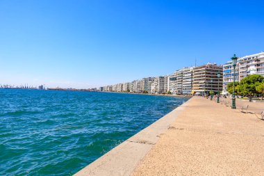 Mavi okyanusu ve binaları olan bir sahil. Binalar yüksek ve balkonları var. Sahil insanlarla dolu ve oturabilecekleri banklar var. 8 Ağustos 2024 Selanik Yunanistan