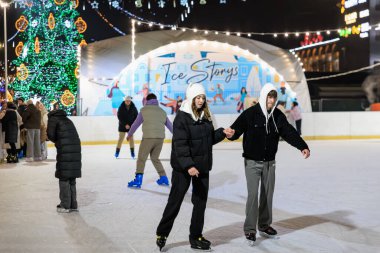Genç yetişkinler gece açık hava buz pateni pistinde festival ışıklarıyla kayıyorlar. 27 Aralık 2024 Balti Moldova.