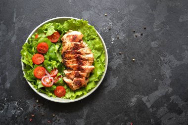 Ispanak ve domatesli ızgara tavuk göğsü salatası. Sağlıklı şekilde kızarmış tavuk göğsü, limon, kiraz domatesleri, ıspanak ve marul yiyor. Karanlık arka plan. Üst Manzara. Metin için boş alan