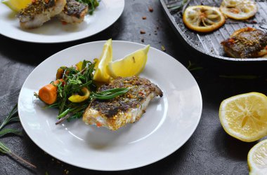 Limonlu ızgara balık, şifalı ot ve biberiye. Beyaz tabakta kızarmış balık filetosu. Güzel bir yemek sunumu. Rucolla salatası ve havuç.