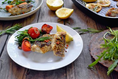 Limonlu ızgara balık, şifalı ot ve biberiye. Beyaz tabakta kızarmış balık filetosu. Güzel bir yemek sunumu. Rucolla salatası ve havuç.