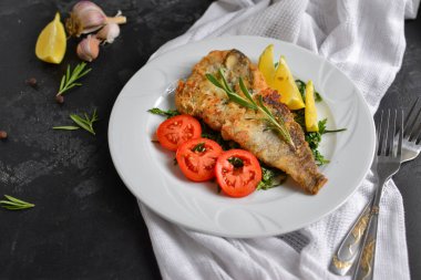 Domatesli, limonlu ve biberiyeli kızarmış balık filetosu. Beyaz tabak. Karanlık arka plan. Beyaz keten peçete..