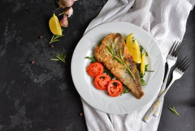 Domatesli, limonlu ve biberiyeli kızarmış balık filetosu. Beyaz tabak. Karanlık arka plan. Beyaz keten peçete. Üst görünüm