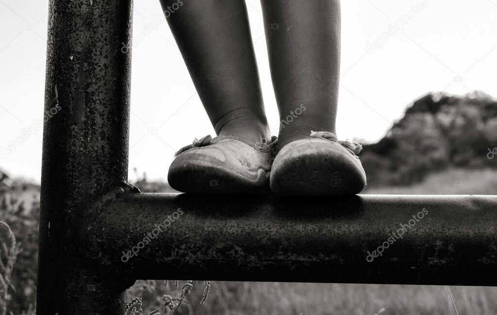Close up black and white photo of a cute caucasian child. Childhood moments. The girl climbs and plays the iron pipe outdoors in the village. Baby feet
