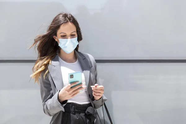 A young woman wearing formal clothes and a medical mask carefully observes her smartphone in the street. Selective focus. Space for text. New normal concept.