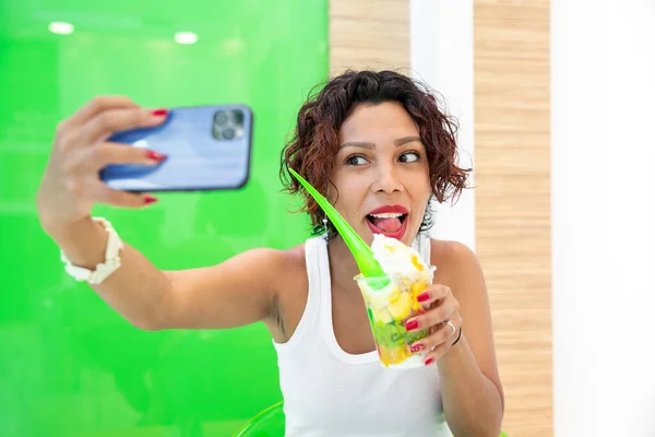 Portrait of a beautiful, brown-skinned woman taking a selfie with her smartphone while holding an ice-cream cup in her hand. Selective focus. Summer and healthy lifestyle concept.
