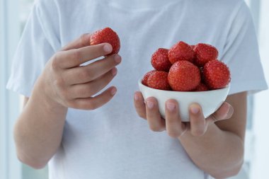 Beyaz gömlekli çocuk bir elinde yuvarlak çilek tabağı, diğer elinde ise yemeye hazır büyük parlak çilek tutuyor. Lezzetli ve vitaminli yiyecekler. Sağlıklı yeme kavramı