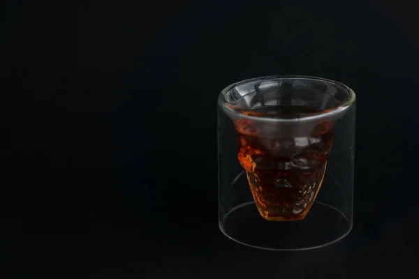 skull shaped glass of brown alcohol drink and on black background. Copy space for text. Poisoned influence of alcohol. Low key image