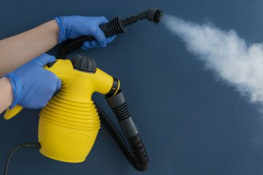 Mavi eldivenli kadın elinin yakın plan görüntüsü. Mavi arka planda beyaz buharlı ev buhar jeneratörü. Alerji, virüs, toz dezenfeksiyonu. Temizlik konsepti.