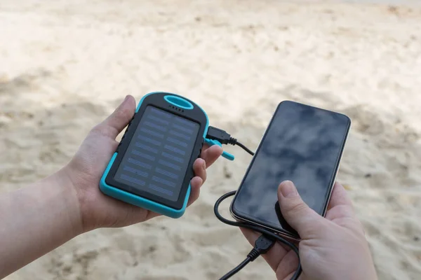 Kadın elleri bir elinde güneş enerjisi, diğer elinde siyah akıllı telefon tutuyor. Plajda elektrik olmadan telefon aküsünü şarj etmeye çalışıyorum. Her durumda temasta kalabilme yeteneği.