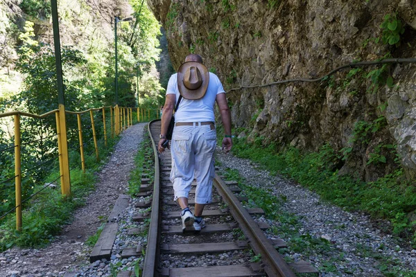 back view of man with straw hat on his back walks on abandoned old railway made in rocks mountains. Traveling by foot concept. Summer vacation in unusual places.