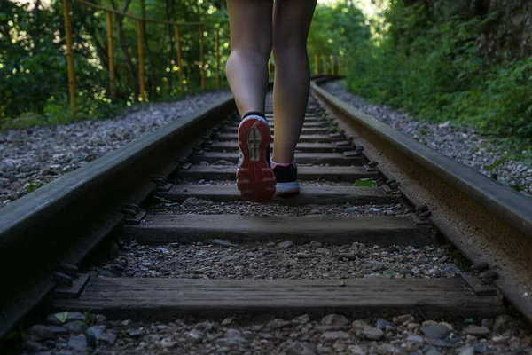 woman is walking on old abandoned rails in green forest, using railway as touristic trail. Summer vacation, traveling by foot concept