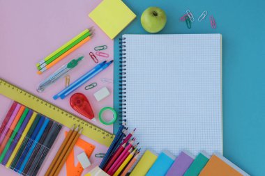 Okul malzemelerinin üst görüntüsü ve yeşil elma mavi ve pembe arka planda yatıyor. Boşluğu beyaz defterde metin için kopyala. Okul konseptine dönelim.. 