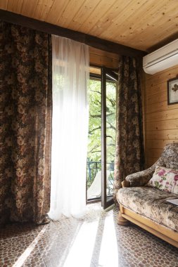 Açık balkon kapısı ve etrafında perdeler olan rahat bir oda. Odanın dışında aydınlık bir yaz günü, evin dışında yeşil park. Rahatlatıcı iç mekan. 