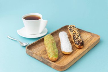 Üzerinde krema olan üç farklı lezzetli eklem tahta tepside yatar, beyaz fincan açık mavi arka planda hamur işine yakındır. Metin için alanı kopyala.