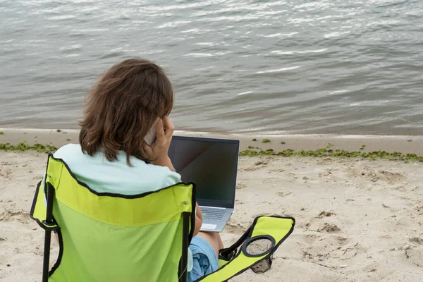 young female sit on bank of river with laptop on knees and smartphone in her hand. Copy space for text. Remote work or education concept.