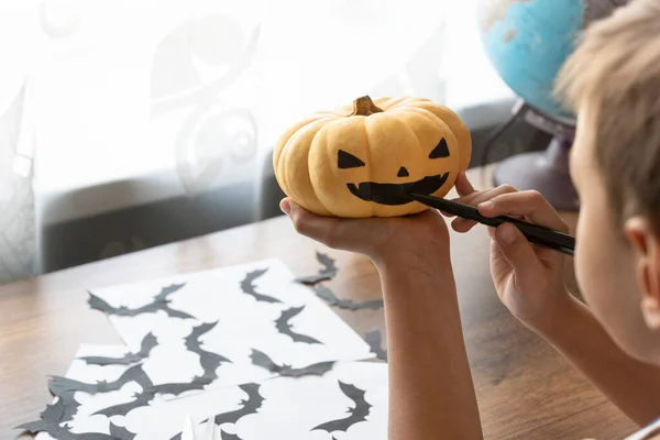 Cadılar bayramı hazırlıkları. Turuncu balkabağına yara izi bırakan bir çocuğun görüntüsü. Masanın yanında siyah yarasa siluetleri var. Cadılar Bayramı konsepti