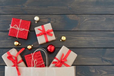 Kırmızı kurdeleyle bağlanmış küçük hediye kutularının olduğu düz bir yatak. Kese kağıdından dışarı bak ve parlak yeni yıl toplarıyla birlikte koyu ahşap arka planda uzan. Yeni yıl alışverişi. Uzayı metin için kopyala. Noel ve yeni yıl konsepti. 