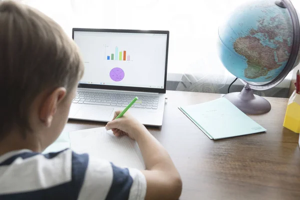 schoolboy is doing online math lesson, writing tasks in notebook from  laptop screen. Globe and copybooks lie near. Home education concept.