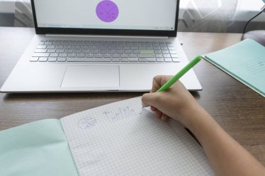 Çocuğun elleri okul defterine matematik ödevi yazıyor, ekranı çizelgeli dizüstü bilgisayar da masanın yanında. Uzaktan eğitim kavramı. Yeni gerçeklik