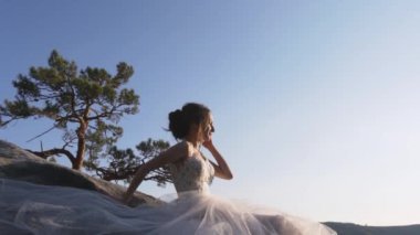 Uzun beyaz, gür bir elbise ve siyah saçlı bej ayakkabılar giyen genç güzel bir kız gün batımında gökyüzüne karşı büyük bir taşın üzerinde oturuyor ve eli taşa yaslanmış ve güneş yüzüne parlıyor.