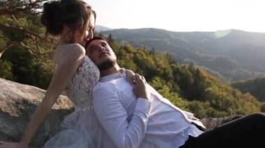 Genç evli bir çift, dağlarda yüksek bir kayanın tepesinde ormanın arka planında ve gün batımında gökyüzünde oturuyor. Adam sırtı kıza dönük ve gözlerinin içine bakıyor.