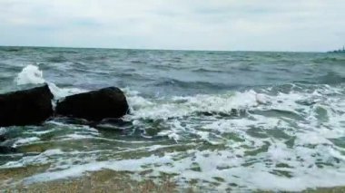 Denizde fırtına, yüksek dalgalar kumlu sahili yıkıyor. Deniz dalgaları taşlara ve köpüğe çarpar. Kıyıdan limana, mavi deniz, mavi gökyüzü.