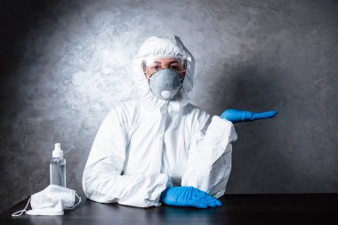 Tıbbi maskeli bir hemşire ve viral koruyucu giysi. Doktor, tıbbi maske ve viral koruyucu giysiyle masada oturuyor. Doktor ilaç ve antiseptiğin yanında oturuyor..