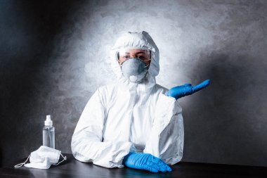 Tıbbi maskeli bir hemşire ve viral koruyucu giysi. Doktor, tıbbi maske ve viral koruyucu giysiyle masada oturuyor. Doktor ilaç ve antiseptiğin yanında oturuyor..