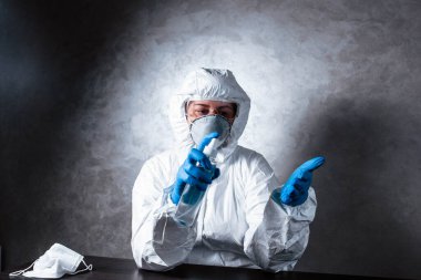 Tıbbi maske takan ve viral koruyucu giysili bir hemşire. Doktor, tıbbi maske ve viral koruyucu giysiyle masada oturuyor. Doktor elleri dezenfekte eder..