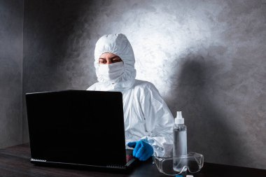Beyaz maske takan ve beyaz koruyucu giysili tıbbi eldiven giyen bir virüs uzmanı dizüstü bilgisayarla çalışıyor ve koronavirüs hakkında bilgi topluyor. İzole edilmiş ve salgın yüzünden panik içinde..