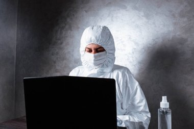 Beyaz maske takan ve beyaz koruyucu giysili tıbbi eldiven giyen bir virüs uzmanı dizüstü bilgisayarla çalışıyor ve koronavirüs hakkında bilgi topluyor. İzole edilmiş ve salgın yüzünden panik içinde..