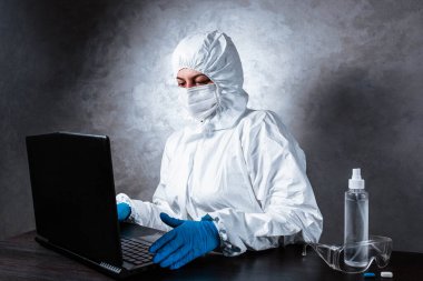 Beyaz maske takan ve beyaz koruyucu giysili tıbbi eldiven giyen bir virüs uzmanı dizüstü bilgisayarla çalışıyor ve koronavirüs hakkında bilgi topluyor. İzole edilmiş ve salgın yüzünden panik içinde..