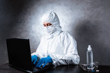 Beyaz maske takan ve beyaz koruyucu giysili tıbbi eldiven giyen bir virüs uzmanı dizüstü bilgisayarla çalışıyor ve koronavirüs hakkında bilgi topluyor. İzole edilmiş ve salgın yüzünden panik içinde..