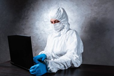 Beyaz maske takan ve beyaz koruyucu giysili tıbbi eldiven giyen bir virüs uzmanı dizüstü bilgisayarla çalışıyor ve koronavirüs hakkında bilgi topluyor. İzole edilmiş ve salgın yüzünden panik içinde..