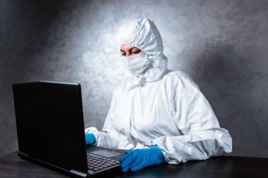 Beyaz maske takan ve beyaz koruyucu giysili tıbbi eldiven giyen bir virüs uzmanı dizüstü bilgisayarla çalışıyor ve koronavirüs hakkında bilgi topluyor. İzole edilmiş ve salgın yüzünden panik içinde..