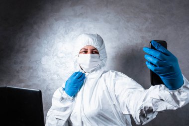 Beyaz maskeli ve tıbbi eldivenli viral koruyucu giysili genç bir kadın görüntülü bir aramada arkadaşlarıyla konuşuyor çünkü doktor koronavirüs salgınının yayılması nedeniyle karantinada..